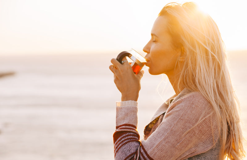 young blonde woman stading on a beach drinking a cup of tea with natural shilajit